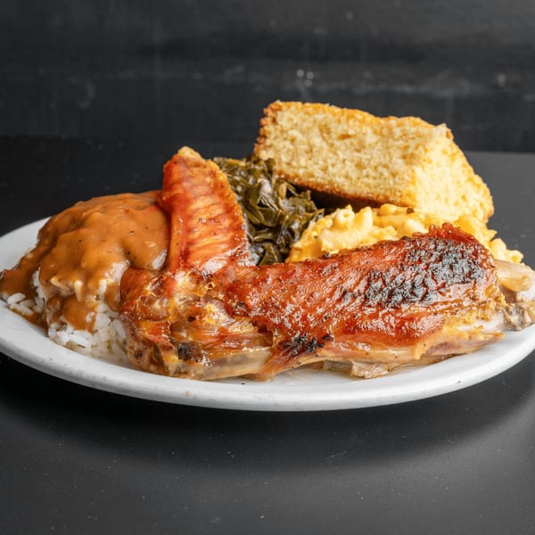 A soul food plate with a roasted turkey wing, rice and gravy, collard greens, mac and cheese, and cornbread.